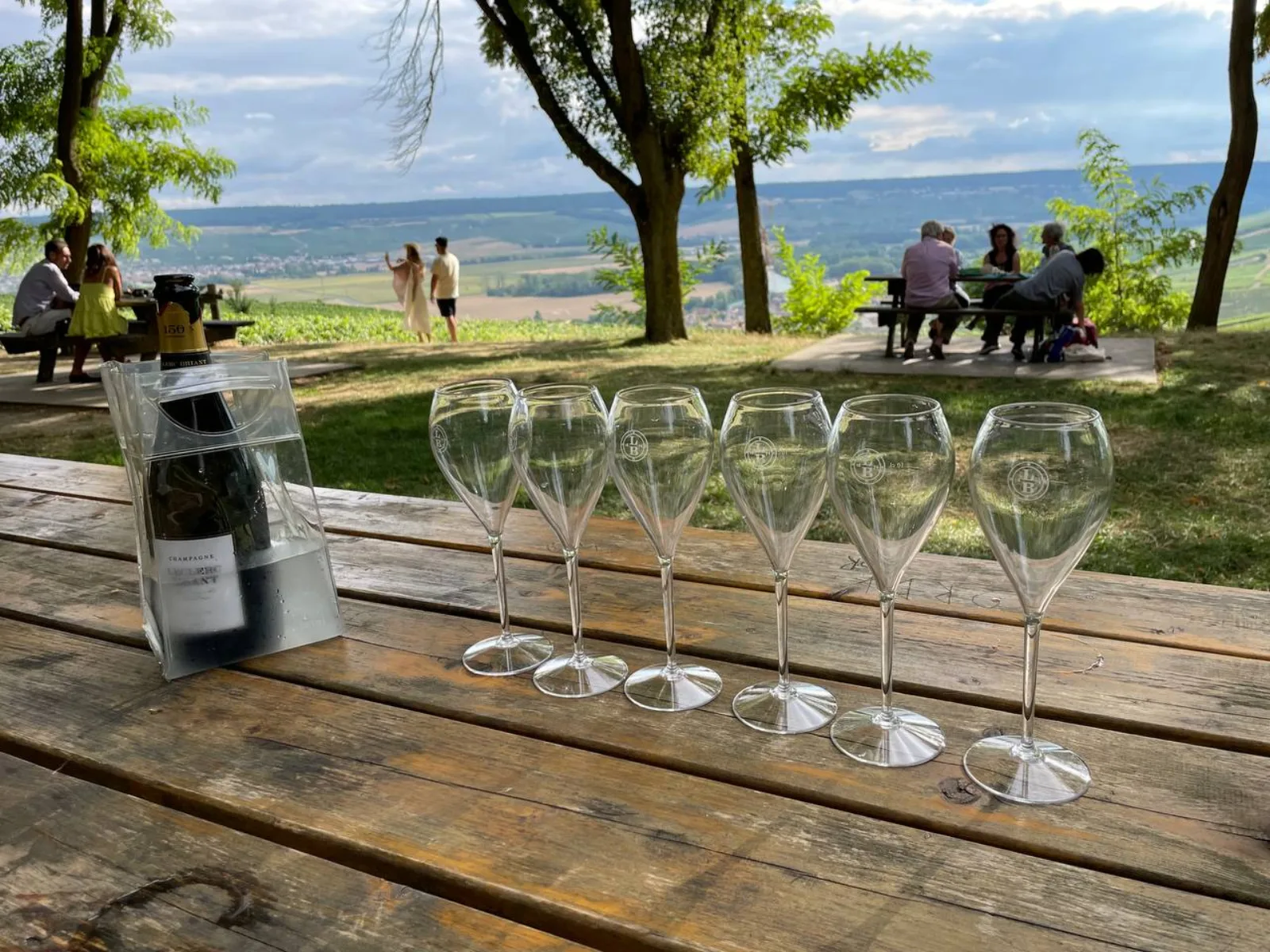 Six empty Champagne flutes and a chilled bottle on a wooden table at a viewpoint