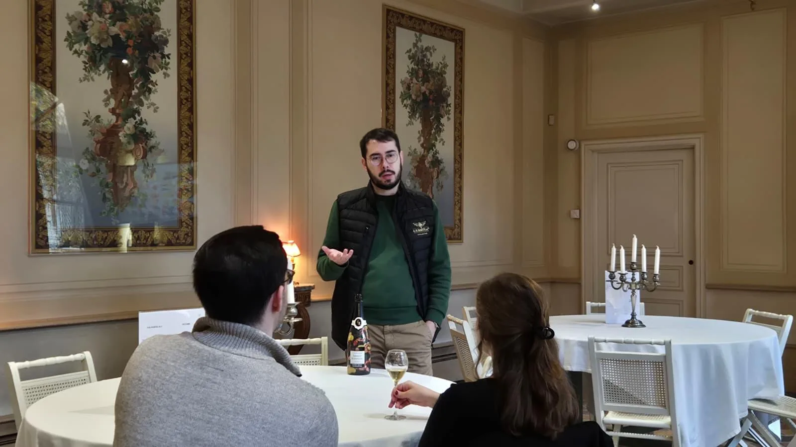 A guide presenting a bottle to seated guests in a tapestried tasting salon